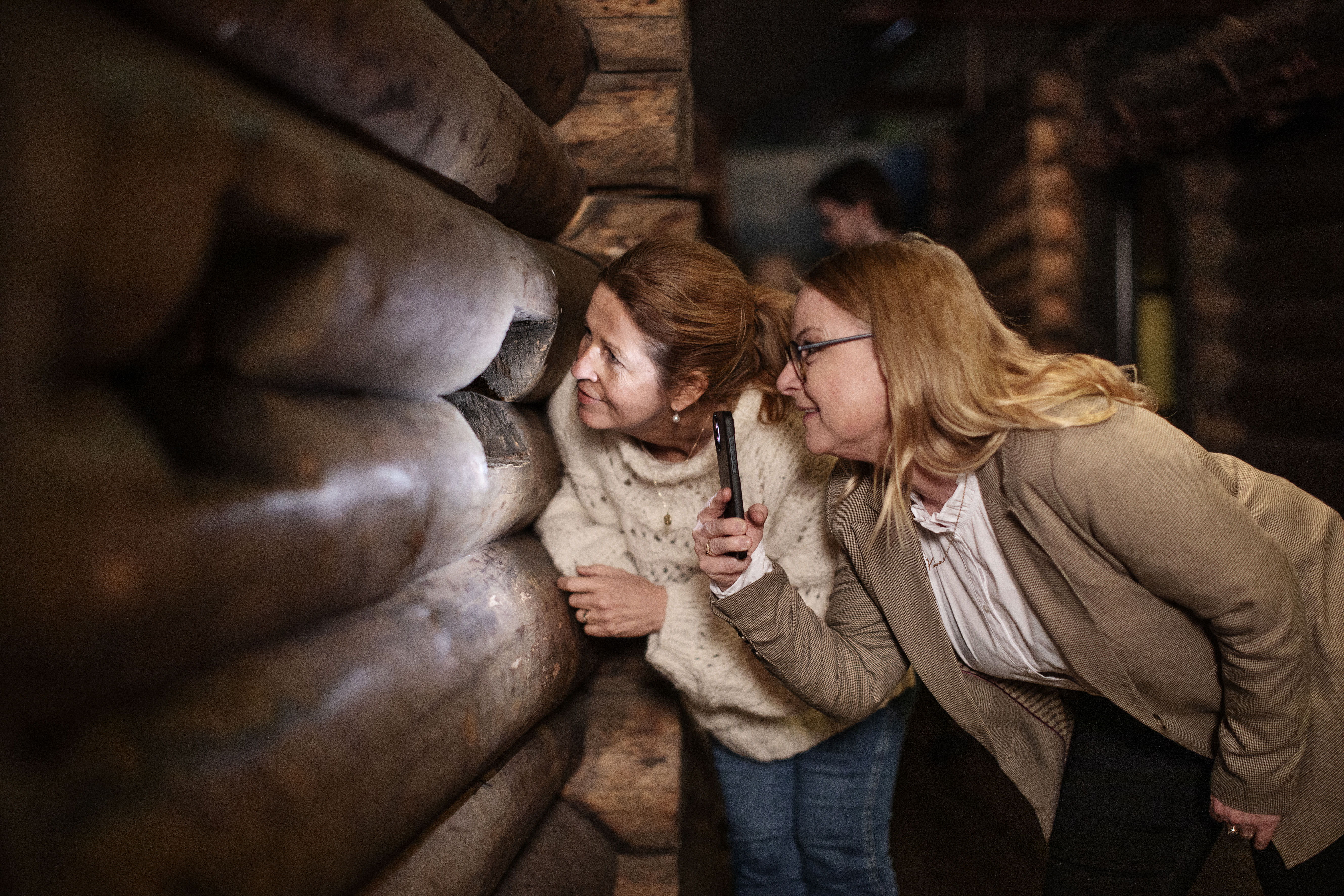 Two women look through an opening in a model of a wooden house. One woman shines a light with her mobile phone.
