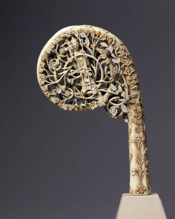A intricately carved crozier made of walrus irvory. The design shows a bishop among detailed greenery. The background is purple-gray.