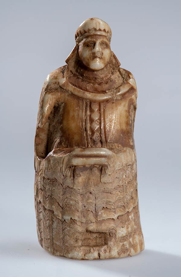 A worn chess piece in the form of a king. He wears a crown and detailed clothing, and sits on a tall throne against a gray background.
