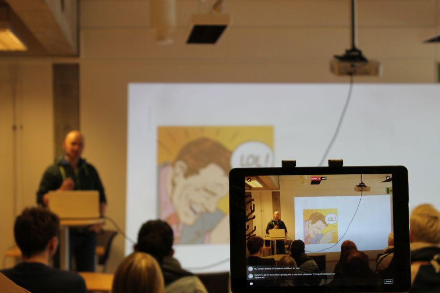 A classroom with a lecturer at the podium,  being recorded by a mobile phone. The audience in chairs looks at a screen with cartoon image. 
