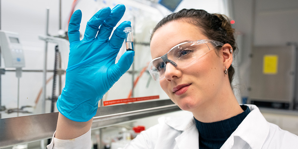 A female student looks at a small glass of chemicals. The student is wearing a white lab coat and protective glasses. Photo