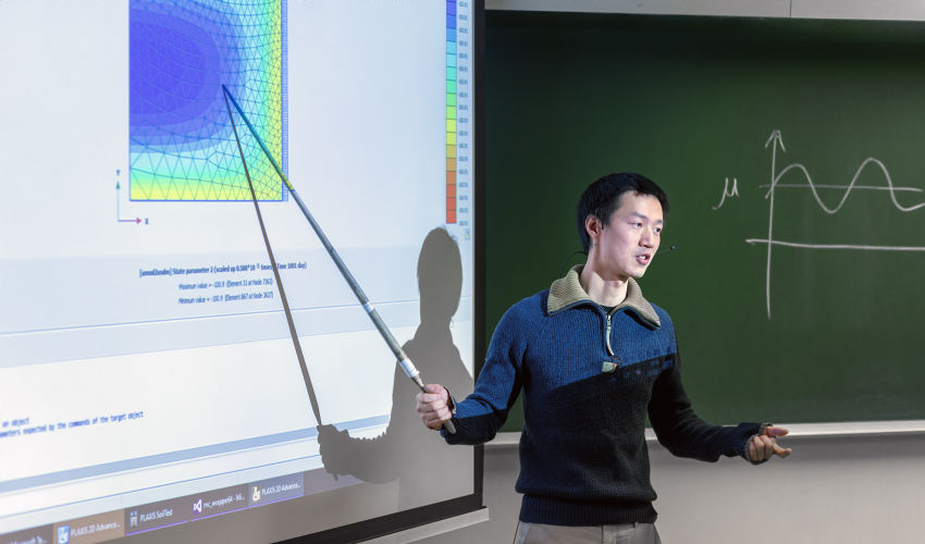 The teacher stands in front of a screen with a presentation and points with a pointer stick.