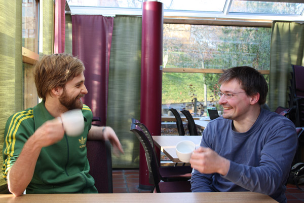 Peter og Frank spiser rundstykker og drikker kaffe på en kafé.