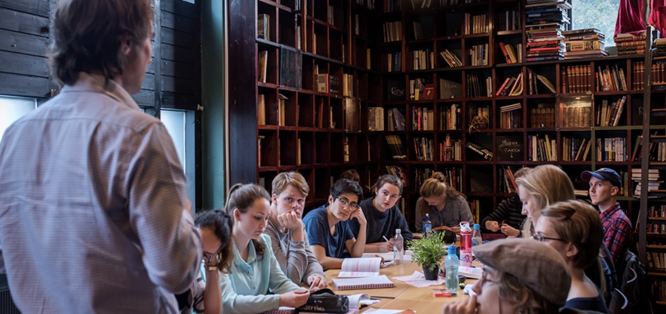 A lecturer in front of students around a table in a library or a room with many books.